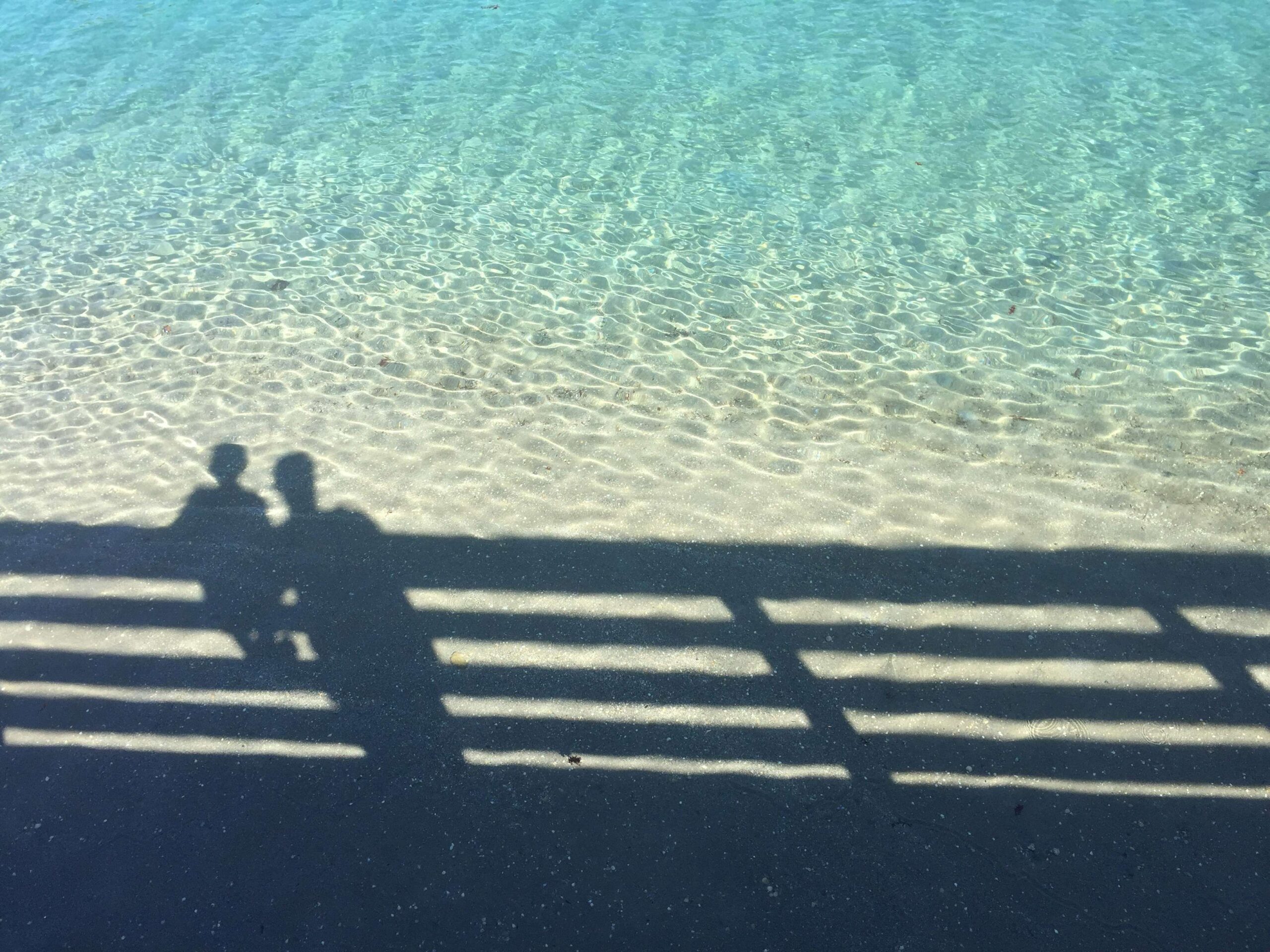 shadow of a parent and child looking over a fence into clear, tranquil sea